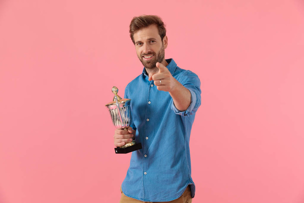 Счастливый случайный парень в джинсовой рубашке, держащий трофей и указывающий пальцем
 - Фото, изображение