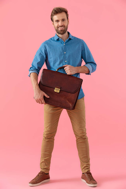 šťastný ležérní chlap v džínové košili s úsměvem a drží kufr - Fotografie, Obrázek