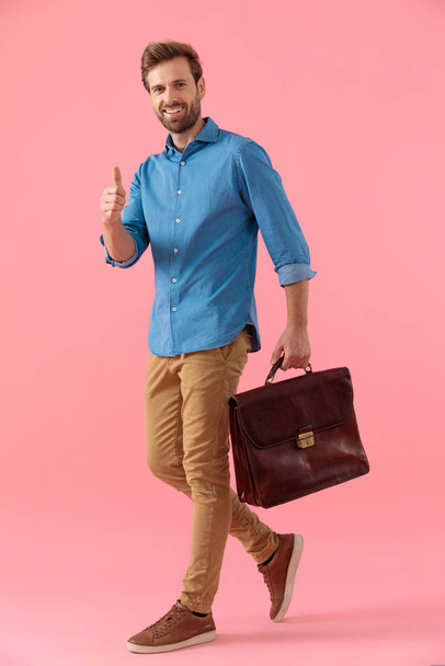 happy casual guy holding suitcase and making thumbs up sign - Photo, Image