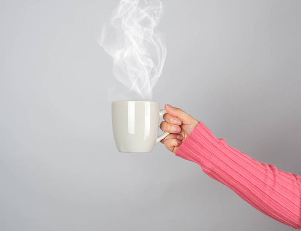 gray ceramic mug in a woman's hand, white steam comes out of the cup, gray background - Foto, imagen