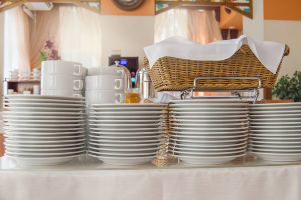 Gros plan d'un groupe de tasses à thé en céramique blanche et assiettes debout sur une table avec une nappe blanche, plats propres dans le restaurant de l'hôtel préparés pour le petit déjeuner des invités
 - Photo, image