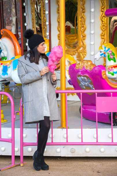 General shot of young brunette woman, with black cap, looking to the right, with pink cotton candy, in front of a carousel, with sunset light, vertically, in Madrid, Spain - Photo, image
