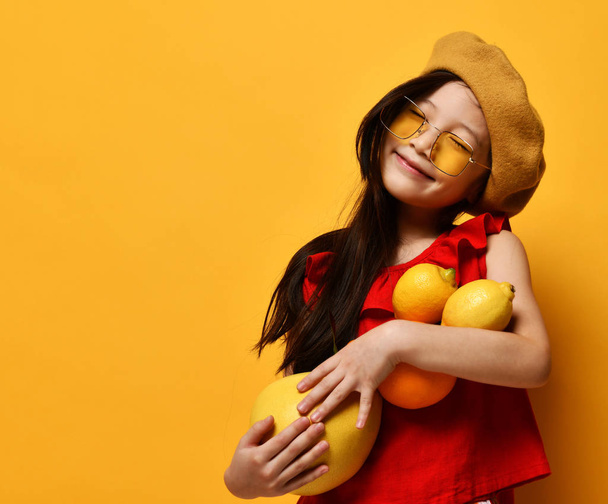 Little asian kid in sunglasses, brown beret, red blouse. Smiling with closed eyes, holding pomelo, orange and lemons. Studio shot. - Фото, изображение