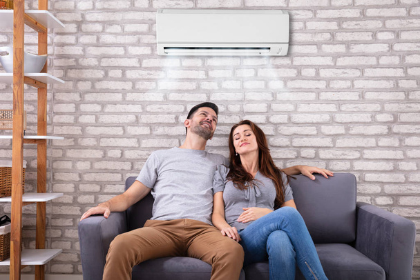 Happy Young Couple Sitting Under Air Conditioner On Couch At Home - Foto, Bild