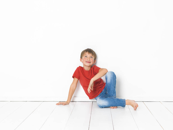 cute and blond boy with red shirt and blue jeans is posing on white wooden floor in front of white background - Foto, Bild