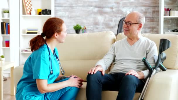 Žena zdravotní sestra mluvit a starat se o starého zdravotně postiženého muže sedí na pohovce - Záběry, video