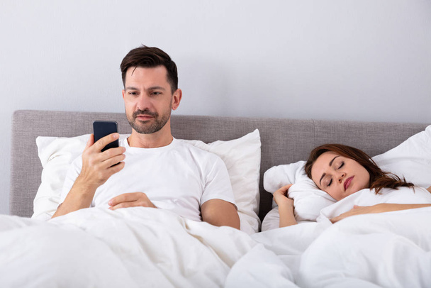 Smiling Man Using Smartphone While His Wife Sleeping On Bed In Bedroom - Фото, изображение