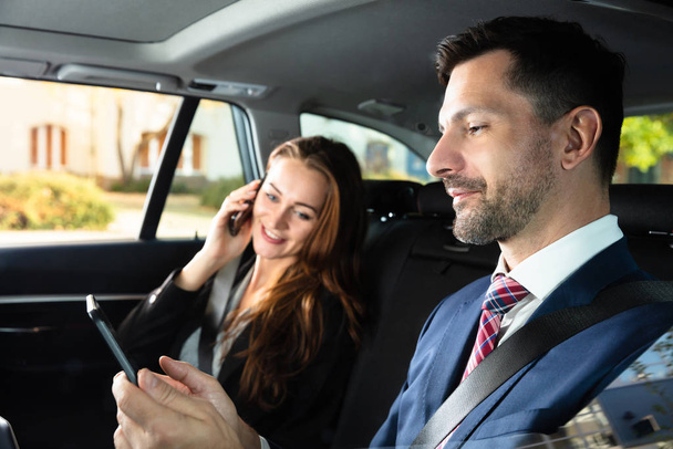 Happy Young Businesswoman Sitting Inside Car Beside Her Male Colleague Using Smartphone - Foto, afbeelding