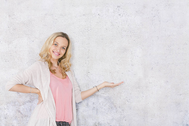 beautiful, blond woman is pointing at and posing in front of grey background - Zdjęcie, obraz