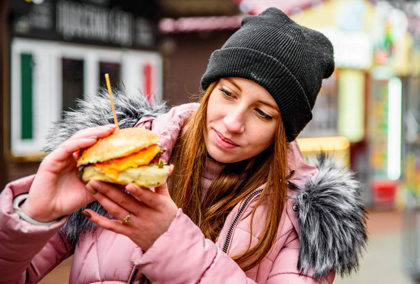 Street food. young woman holding juicy burger and eating oudoor winter - Fotó, kép