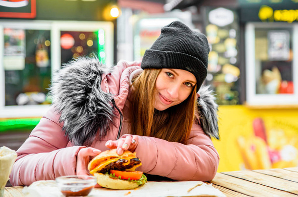 Street food. young woman holding juicy burger and eating oudoor winter - Photo, image