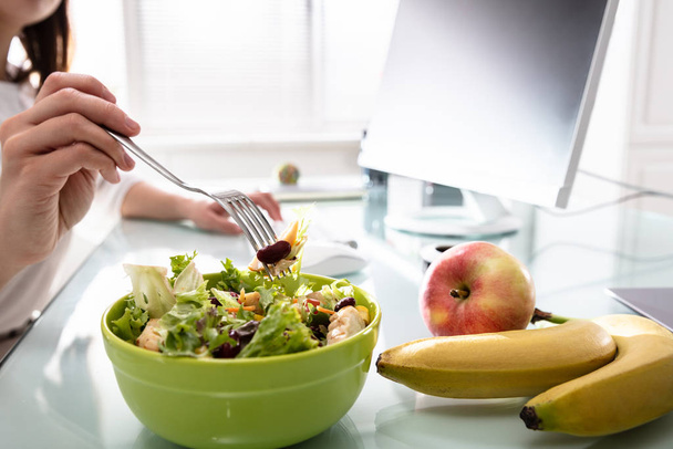 Close-up Of Woman's Hand Holding Fork While Having Healthy Salad On Office Desk - Fotografie, Obrázek