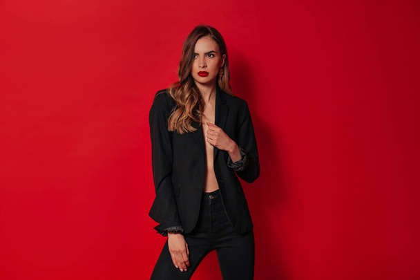 Elegante mujer confiada en traje negro posando sobre fondo rojo en el estudio
  - Foto, Imagen