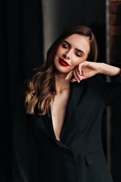Studio portrait of pretty woman with red lips and wavy hair wearing black jacket posing at camera during photoshoot and touching her face - Foto, afbeelding