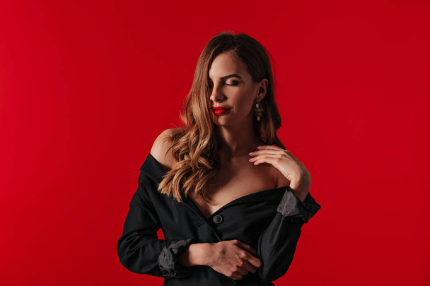 Привлекательная стильная женщина с красными губами и волнистыми волосами в черном платье с обнаженными плечами, позирующая с закрытыми глазами и улыбающаяся на красном фоне
  - Фото, изображение