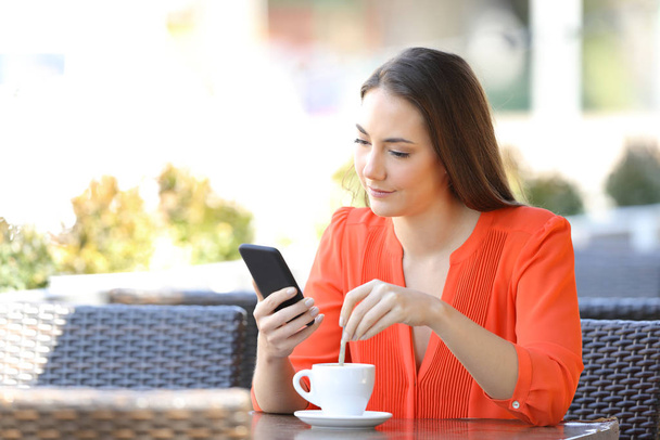 Woman stirring coffee checking smart phone content sitting in a bar terrace - Fotó, kép