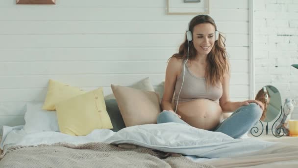 Lächelnde werdende Mutter mit Kopfhörern auf nacktem Bauch im Schlafzimmer. - Filmmaterial, Video