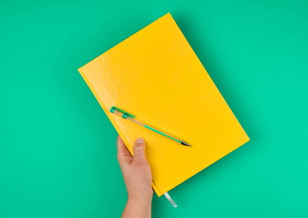 hand holds large closed yellow notebook and green pen, top view - Foto, afbeelding