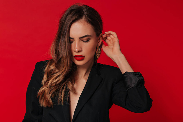 Sensual pretty woman with red lips wearing gold earrings and black jacket posing over red background looking down and straightens hair - Photo, Image