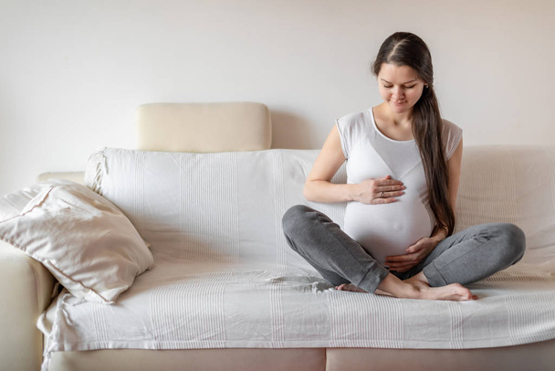 Young pregnant woman sitting on white sofa and keeping hands on belly - Foto, Bild