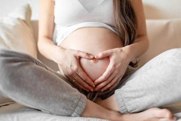 Mujer embarazada joven sentada en un sofá blanco y manteniendo las manos en el vientre
 - Foto, Imagen