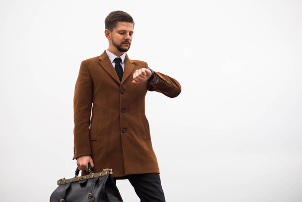 Portrait d'un jeune homme de trente ans, dans un style d'affaires, avec un sac en cuir à la main, regardant sa montre. Contre le ciel, style de rue
 - Photo, image