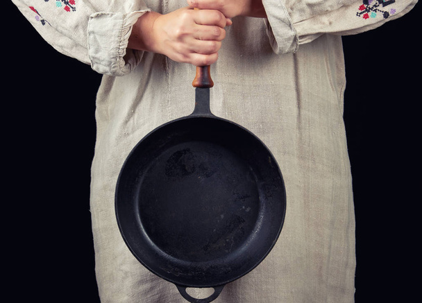 жінка в сірій лляній сукні тримає з двома руками круглу чавунну сковороду, чорний фон
 - Фото, зображення