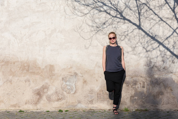 Modern trendy fashionable woman wearing sunglasses leaning against old textured retro wall with tree shadow falling on the wall. Graphical and textured artisic image. - Fotografie, Obrázek