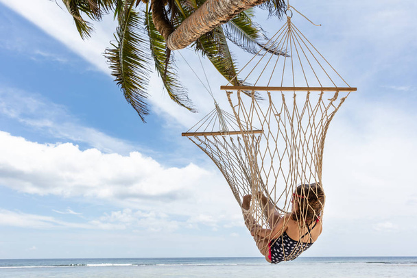 Uklidňující dívka v houpací síti zavěšená na palmových stromech u moře proti modrému nebi - Fotografie, Obrázek