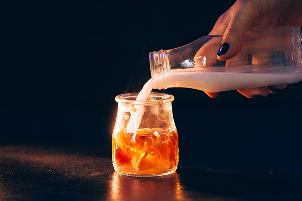 Tyttö kaataa maitoa lasipullosta lasipurkkiin teetä mustalla taustalla lämpimällä valolla
 - Valokuva, kuva