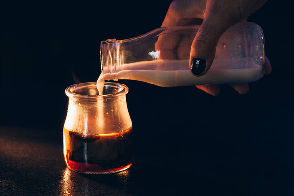 Menina derrama leite de uma garrafa em um frasco de café em um fundo escuro com luz quente
 - Foto, Imagem