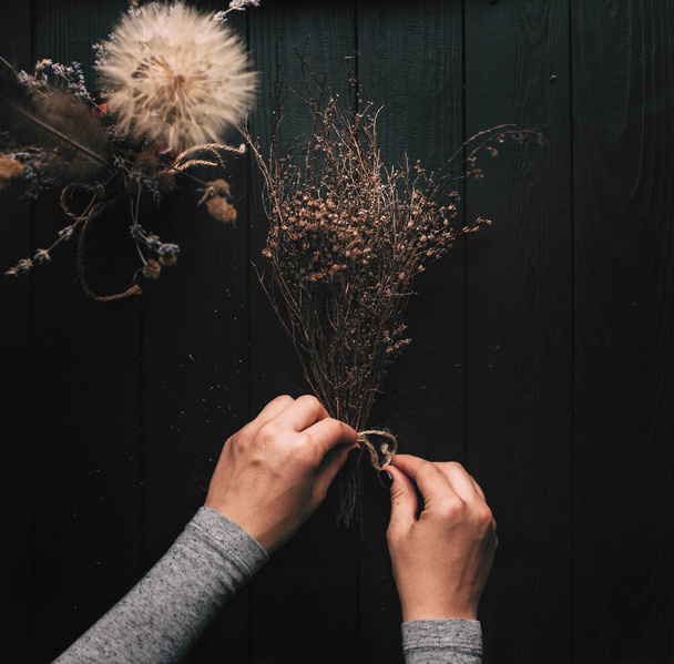 Creating a bouquet of dried herbs on a dark wooden background. Hands in the frame. - Фото, изображение