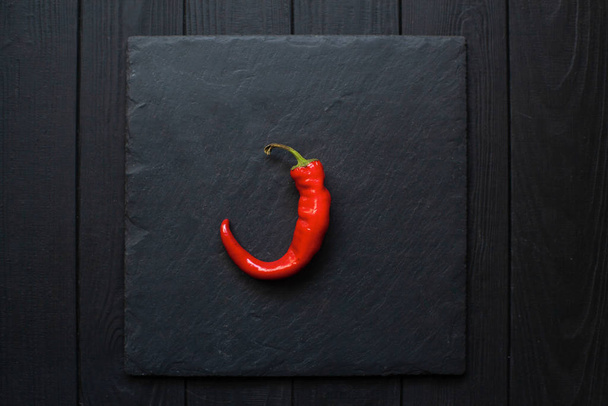 Ζεστό κόκκινο πιπέρι σε ένα πίνακα πλάκα που βρίσκεται σε μαύρο ματ φόντο ξύλο - Φωτογραφία, εικόνα