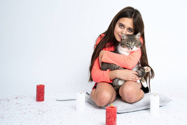 Little brunette girl sitting on the pillow, hugging a cat and looking at the camera, red and white candles are placed around, isolated over white background - Foto, afbeelding