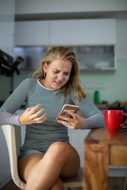 Teenage girl using her cell phone, being angry, while having a cup of tea in modern kitchen setting (shalllow DOF, color toned image) - Foto, Imagem