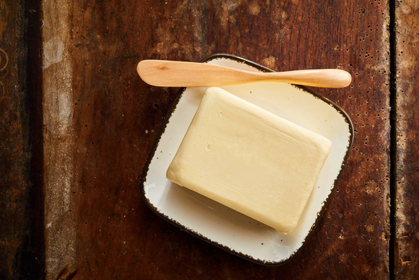 Whole pat of farm fresh creamy butter on a small rectangular butter dish with wooden spreader or knife viewed top down on a rustic wood table - Photo, Image