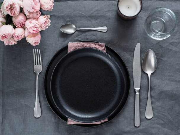 Beautiful table setting on gray linen tablecloth. Festive table setting for wedding dinner with pink spray roses and scented cabdle. Holiday dinner with black plates. Dinner, utensil. Copy space - Fotografie, Obrázek