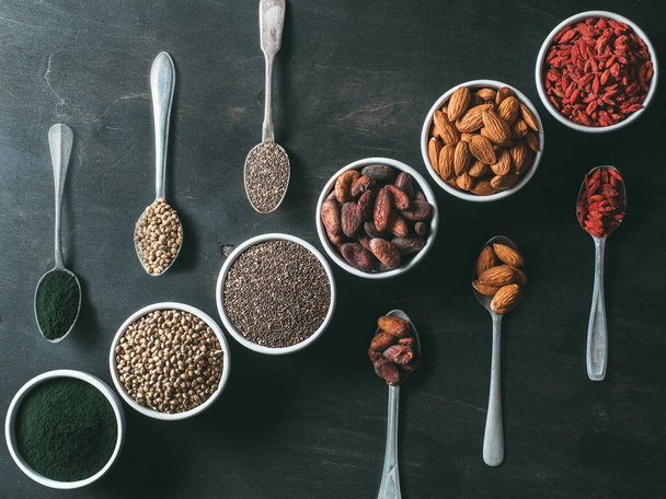 Above view of various superfoods in spoons and small bowl on black wooden table background. Superfood close up chia seeds, spirulina, cocoa bean, goji berry, hemp seeds, almond. Top view, flat-lay - Photo, image