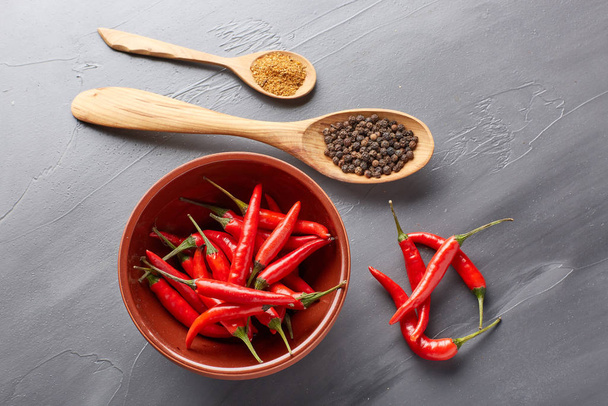 Still life with different peppers (whole black dry and powdered mix peppers in a wooden spoons, fresh red chile peppers in a bowl) on a gray background. Top view. - Фото, изображение