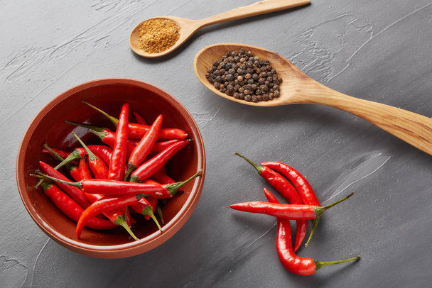 Still life with different peppers (whole black dry and powdered mix peppers in a wooden spoons, fresh red chile peppers in a bowl) on a gray background. Top view. - Foto, imagen