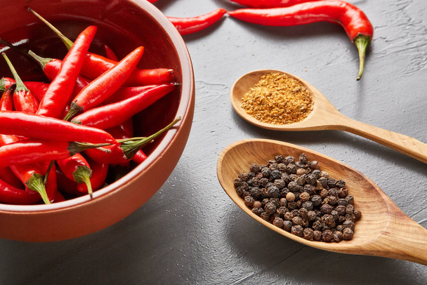 Still life with different peppers (whole black dry and powdered mix peppers in a wooden spoons, fresh red chile peppers in a bowl) on a gray background. Top view. - Photo, Image
