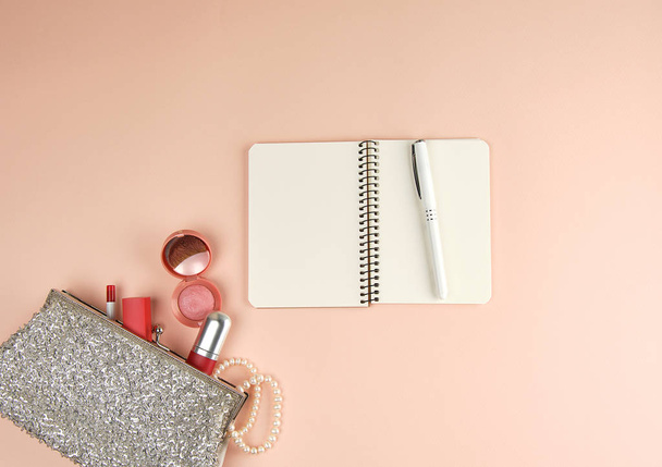 open notebook with clean white sheets and a pen, next to an open women's handbag with cosmetics on a peach background, top view - Foto, Bild