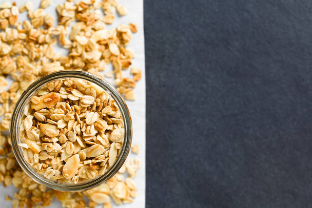 Homemade baked crunchy oatmeal, sliced almond, honey and coconut oil breakfast granola in glass jar, photographed overhead on slate with copy space on the right (Selective Focus, Focus on the granola in the jar) - Photo, image