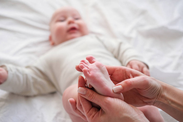 Baby feet in mother hands. Young caucasian woman makes massage for happy infant baby on white bed at home. Babycare, sport and happy motherhood. - Foto, immagini