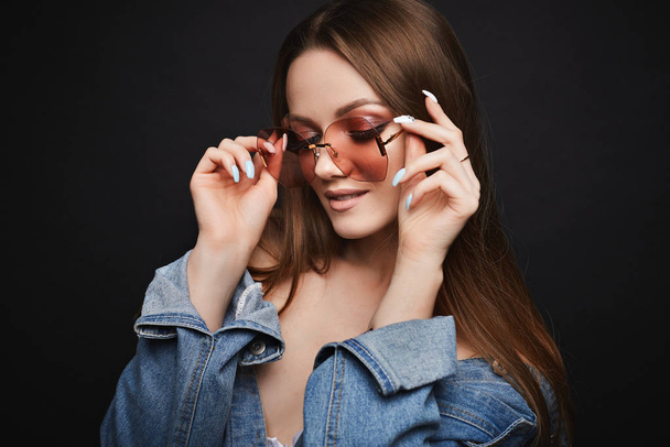 Young model woman in cool pink sunglasses and denim jacket posing on black isolated background. Attractive girl with bright makeup in jeans jacket smiling and posing in studio. Accessories and makeup - Foto, Imagem
