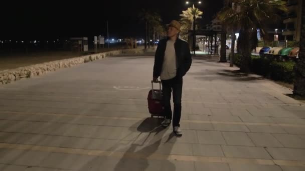 Un viajero con sombrero y chaqueta con una maleta roja camina bajo las palmeras a lo largo del paseo nocturno
 - Metraje, vídeo