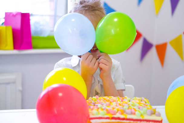 Birthday boy hid behind colorful balloons near birthday rainbow cake. Festivel background. Funny birthday party - Фото, изображение