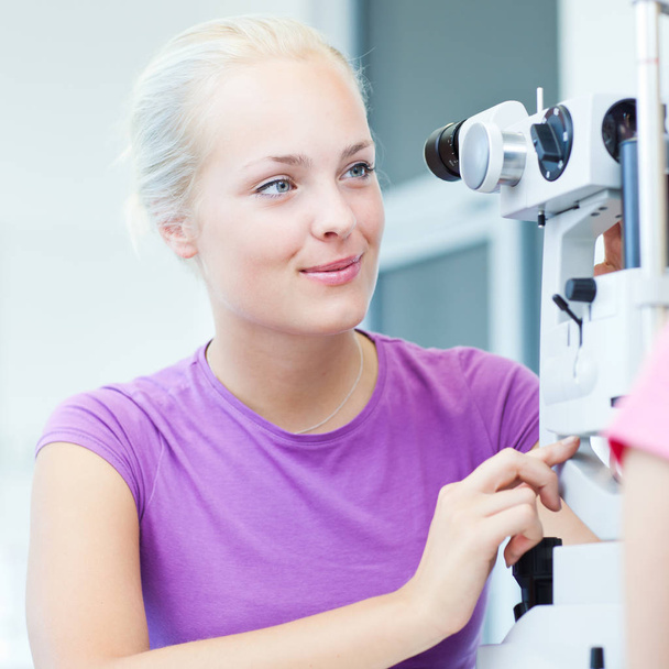 optometry concept - portrait of a pretty young optometrist using a slit lamp to check her patient&#039 - Foto, afbeelding