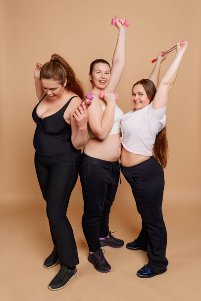 Three fat woman posing with fitness accessories on beige background - Foto, imagen