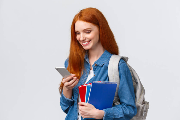 Schöne gut aussehende Frau mit roten Haaren, auf dem Weg zum College, mit Rucksack, Notizbücher lächelnd als Antwort Freund Nachricht, Chat Blick mobilen Bildschirm erfreut, weißer Hintergrund - Foto, Bild